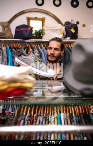 Bel giovane uomo che guarda le scarpe mentre sceglie nuove sneakers in un piccolo negozio di abbigliamento Foto Stock