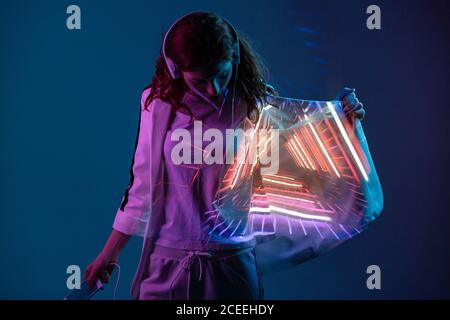 Giovane donna in cuffie guardando la proiezione di luce al neon sul blazer in studio Foto Stock