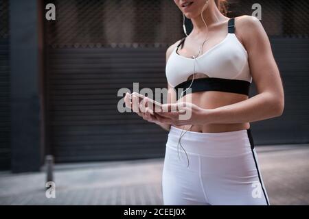 Foto senza volto di Muscular Woman in vestito sportivo bianco con cuffie e smartphone per strada Foto Stock