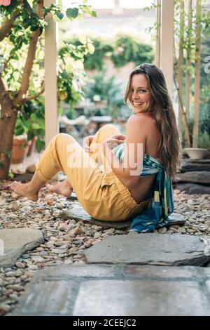 Vista laterale della bella donna a piedi nudi che ride e guarda macchina fotografica mentre si siede su un sentiero in pietra in serra leggera Foto Stock