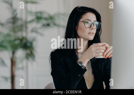 Foto di concentrato brunette femmina copywriter bevande caffè, lavori su attività creativa, indossa spettacoli e costume nero formale, orologi webinar a. Foto Stock