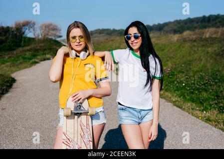 Ragazze teen alla moda in shorts e t-shirt in piedi con tavola lunga sulla strada in campagna?looking macchina fotografica alla luce del sole. Foto Stock