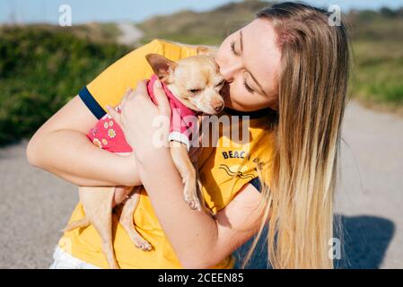 Felice ragazza casual con lunghi capelli biondi che tengono e baciano il piccolo cane Chihuahua in piedi sulla strada sotto il sole. Foto Stock