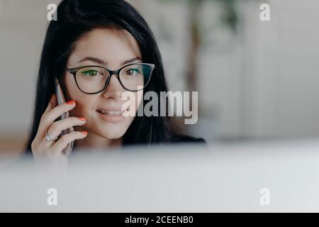 Immagine orizzontale di attraente dipendente donna brunette con espressione concentrata del volto, parla sul telefono cellulare, ha conversazione cellulare, indossa specifiche Foto Stock