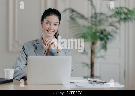 Affascinante donna d'affari di stile lavora freelance al computer portatile, utilizza connessione internet ad alta velocità, tiene occhiali in mano, gode remo preferito Foto Stock