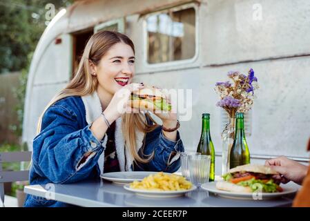 Bella giovane femmina sorridente e mangiare delizioso hamburger mentre si ha cena romantica con un ragazzo irriconoscibile vicino a un vecchio camper Foto Stock