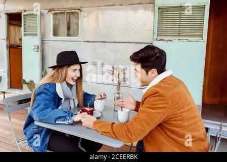 Giovane uomo e donna sorridente e guardandosi mentre si siede al tavolo con bevanda calda e pezzo di torta di compleanno vicino al camper stagionato Foto Stock