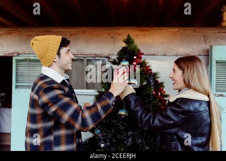 Giovane uomo e donna in abiti alla moda che decorano l'albero di Natale vicino a carovana invecchiata in campagna Foto Stock
