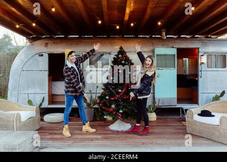 Attraente giovane uomo e donna sorridente e guardando la macchina fotografica mentre mostra splendidamente decorato albero di Natale vicino a furgone shabby Foto Stock