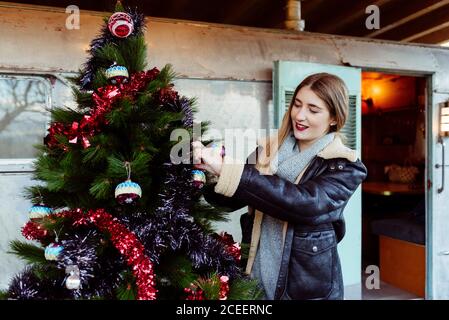 Giovane donna e donna in abiti alla moda decorando l'albero di Natale vicino a carovana invecchiata in campagna Foto Stock