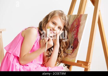 Ritratto di una bella bambina che dipinge una foto in uno studio o in una scuola d'arte. Creativo pittore pensivo bambino dipinge un'immagine colorata su tela con Foto Stock