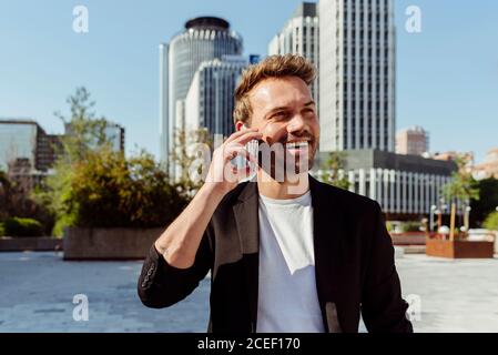Un giovane attraente che sorride con lo smartphone mentre si trova in strada di città moderna in giornata di sole Foto Stock