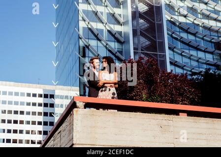 Bell'uomo caucasico sorridente e abbracciante Afro-American Woman dal retro mentre si trova vicino al confine sulla strada della città insieme Foto Stock