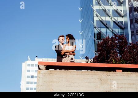 Bell'uomo caucasico sorridente e abbracciante Afro-American Woman dal retro mentre si trova vicino al confine sulla strada della città insieme Foto Stock