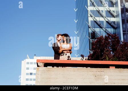 Bell'uomo caucasico sorridente e abbracciante Afro-American Woman dal retro mentre si trova vicino al confine sulla strada della città insieme Foto Stock