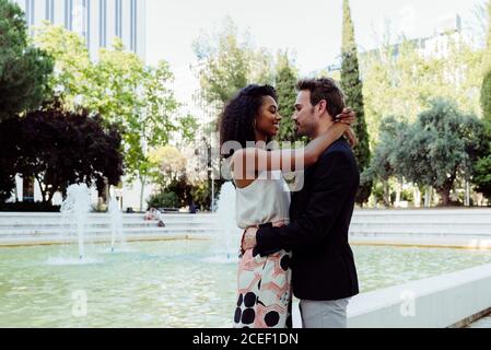 Vista laterale dell'affascinante donna nera e del bell'uomo caucasico che si abbracciano mentre si trova vicino alla bellissima fontana nel parco Foto Stock