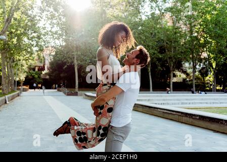 Vista laterale del bell'uomo caucasico che solleva una donna afroamericana ridente mentre si trova in piedi sul vicolo del parco nelle giornate di sole Foto Stock