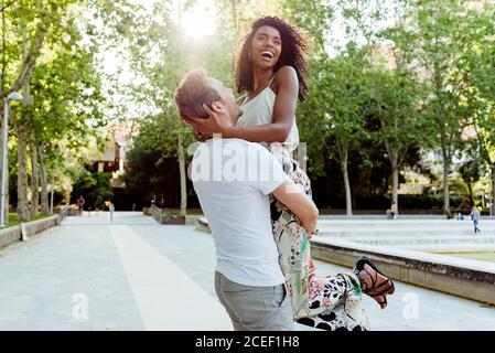 Vista laterale del bell'uomo caucasico che solleva una donna afroamericana ridente mentre si trova in piedi sul vicolo del parco nelle giornate di sole Foto Stock