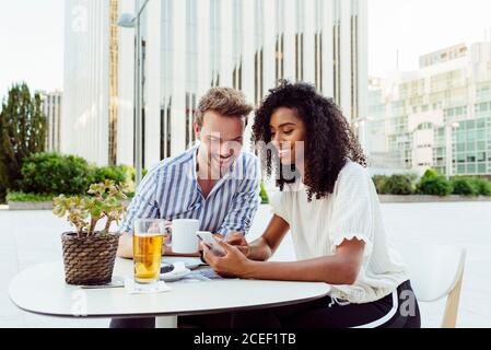 Simpatica coppia multirazziale sorridente e di navigazione smartphone moderno mentre si è seduti al tavolo c'è un bel caffè all'aperto insieme Foto Stock