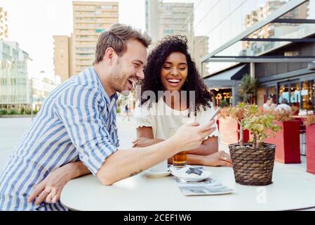 Simpatica coppia multirazziale sorridente e di navigazione smartphone moderno mentre si è seduti al tavolo c'è un bel caffè all'aperto insieme Foto Stock