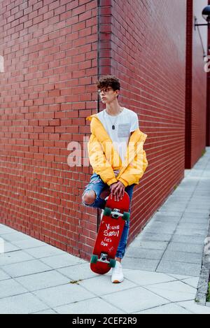 Ragazzo adolescente in occhiali con skateboard poggiato su un angolo sulla strada Foto Stock