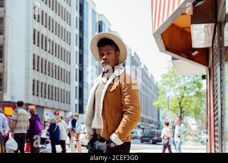 Giovane africano maschio in cappello e zaino guardando su strada con auto in metropoli Foto Stock