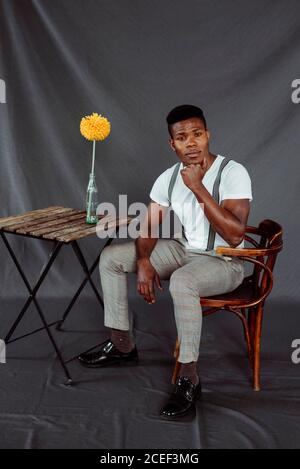 Bell'uomo afro-americano in outfit elegante guardando la macchina fotografica e. seduta su sedia retrò vicino a piccolo tavolo con fiore Foto Stock