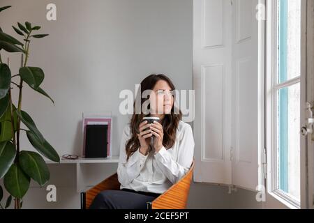 Bella femmina in abito elegante che tiene una tazza di fresco caldo bevete e guardate fuori dalla finestra mentre sedete su una comoda sedia in una camera accogliente Foto Stock
