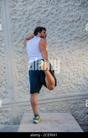 Vista posteriore del bell'uomo adulto appoggiato su una parete di cemento e facendo l'esercitazione di riscaldamento per le gambe durante l'allenamento all'aperto sulla strada Foto Stock