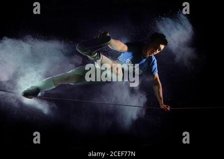 Il liner maschio si bilancia su una linea slackline tenendo un piede a lato in un'esplosione di polvere bianca dipingere con uno sfondo nero Foto Stock