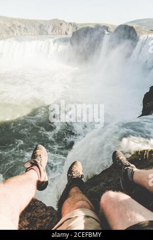 Ritaglio di gambe di viaggiatori in stivali seduti sulla scogliera sopra la maestosa cascata in foschia e luce solare Foto Stock