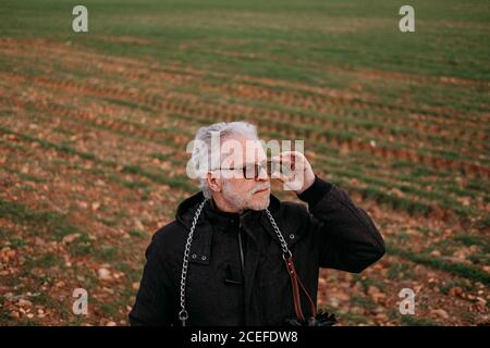 Anziano uomo serio in occhiali da sole e giacca nera guardando via con fiducia sullo sfondo del campo. Foto Stock
