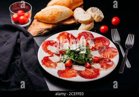 Gustose e sottili fette di carne a strappo cosparse di pomodori tritati e le alette sono fissate su piastra in ceramica bianca con forcelle intorno su tavolo di legno nero dall'alto Foto Stock