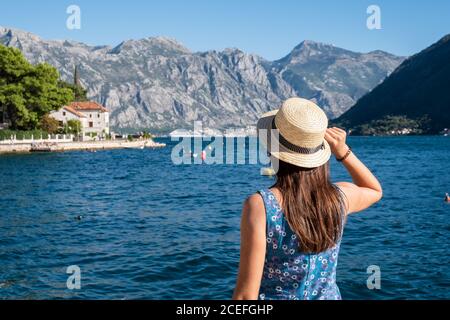Vista posteriore di una donna irriconoscibile in un elegante cappello che guarda bel mare increspato e magnifica cresta di montagna in giornata di sole sul resort Foto Stock
