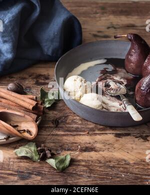 Bastoncini di cannella aromatica giacenti sul piano portapaziente di legno vicino alla ciotola con fusione di gelato e deliziose pere nel vino Foto Stock
