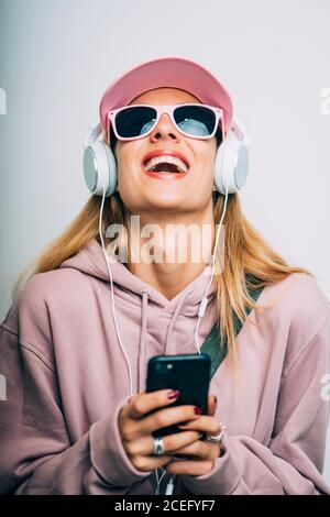 Giovane donna alla moda in pullover e occhiali da sole che ascolta musica in cuffia bianca con telefono cellulare in mano su sfondo bianco Foto Stock
