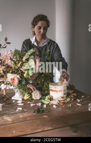 Vista laterale della signora posizionando il piatto con una gustosa torta decorata bloom bud sul tavolo di legno con mazzo di crisantemi, rose e rametti di piante in vaso tra Foto Stock