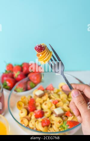 minuscoli pancake su una forchetta con fragole fresche, banana, cioccolato, patatine di cocco e miele in un piatto su sfondo blu. tendenza alimentare 2020. Foto Stock