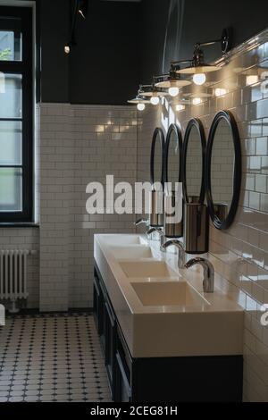 Interni di design elegante di toilette pubblica con bianco pareti piastrellate e lavandini in ceramica sotto luci incandescenti Foto Stock