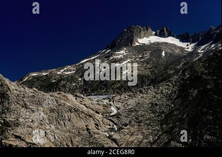 Il ghiacciaio Rhône scorre a circa 30 - 40 m (100 - 130 piedi) all'anno dai campi innevati delle Alpi Urner fino a un punto in cui l'acqua di fusione forma un piccolo lago sotto il Passo Furka nel Cantone Vallese, Svizzera. Da qui, ad un'altitudine di circa 2,208 m (7,244 piedi), un torrente costante si aggita nel bacino della valle molto in basso per iniziare il suo viaggio di 813 km (505 mi), come il fiume Rhône, attraverso la Francia meridionale fino al delta della Camargue, dove scorre nel Mediterraneo. Foto Stock