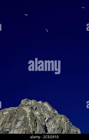 Tre parapendio navigano sulle alte correnti termali in un cielo blu profondo di luglio su una dorsale frastagliata nelle Alpi Urner che si erge sopra il ghiacciaio Rhône di 7.65 km (4.75 mi) nel Cantone Vallese, Svizzera. Un quarto aviatore solista scula la sua superficie robusta. Il ghiaccio del ghiacciaio si scioglie a un'altitudine di circa 2,208 m o 7,244 piedi, con l'acqua di fusione che cade in una profonda conca a valle molto al di sotto per iniziare il viaggio del fiume Rhône di 813 km (505-mi) verso il Mar Mediterraneo attraverso la Francia meridionale e il delta della Camargue. Foto Stock