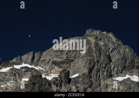 Un parapendio solista appeso in un cielo blu profondo galleggia per sciare la superficie aspro di una dorsale frastagliata nelle Alpi Urner, nel Canton Vallese, in Svizzera, alta sopra il ghiacciaio Rhône di 7.65 km (4.75 mi). Il ghiaccio del ghiacciaio si scioglie a un'altitudine di circa 2,208 m o 7,244 piedi, con l'acqua di fusione che cade in una profonda conca a valle molto al di sotto per iniziare il viaggio del fiume Rhône di 813 km (505-mi) verso il Mar Mediterraneo attraverso la Francia meridionale e il delta della Camargue. Foto Stock
