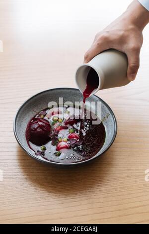 Vista del raccolto della mano maschile che versa succo di barbabietola rosso scuro Dalla caraffa alla ciotola grigia con verdure nordiche tritate e. potherbs posto su tavolo di legno chiaro Foto Stock