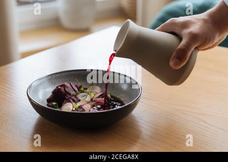 Vista del raccolto della mano maschile che versa succo di barbabietola rosso scuro Dalla caraffa alla ciotola grigia con verdure nordiche tritate e. potherbs posto su tavolo di legno chiaro Foto Stock