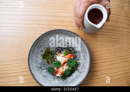 Vista di raccolto della mano maschio che versa salsa spessa rossa da caraffa nel recipiente con insalata tritata con verdure e pesce servita su tavolo di legno Foto Stock