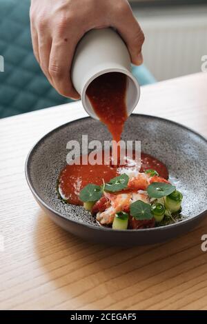 Vista di raccolto della mano maschio che versa salsa spessa rossa da caraffa nel recipiente con insalata tritata con verdure e pesce servita su tavolo di legno Foto Stock