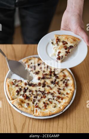 Raccolto colpo da sopra di persona che serve fetta di cotto Deliziosa pizza in stile nordico in piedi al tavolo di legno Foto Stock