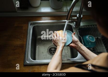 Vista posteriore del raccolto del piattino blu di Woman Hands?cleaning con spugna per il lavaggio dei piatti nel lavandino sul bancone di legno con davanzale con pentole nelle vicinanze Foto Stock