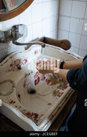 Le mani di pulizia femmina del raccolto sotto che corrono dall'acqua del rubinetto di metallo in vecchio lavello bianco fessurato con ornamento floreale Foto Stock