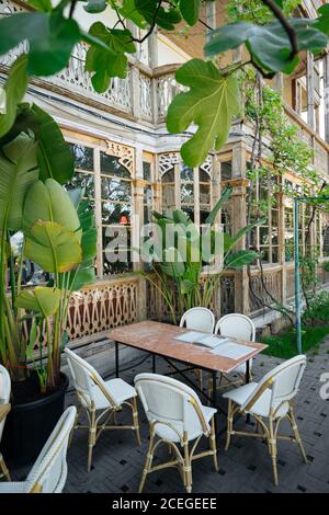 Bel tavolo da caffe' in legno shabby con menu' con sedie in rattan intorno in piedi al vecchio edificio sul marciapiede di strada con piante con foglie grandi intorno Foto Stock